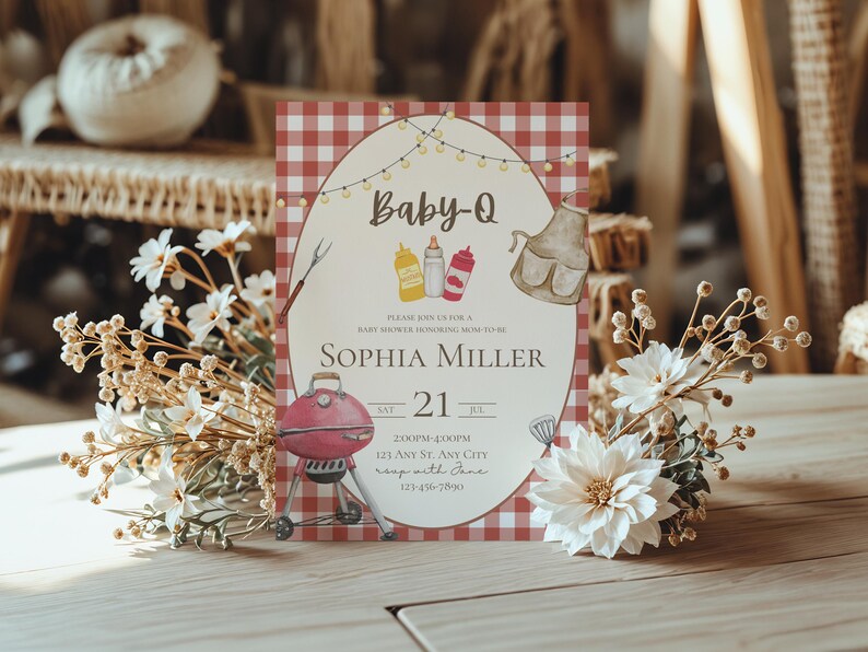 Op de afbeelding: Baby-Q babyshower uitnodiging met een rood-witte geruite achtergrond. De uitnodiging bevat een barbecue-illustratie, tekst en decoratieve elementen. Witte bloemen en gedroogde bloemaccenten omringen de uitnodiging.