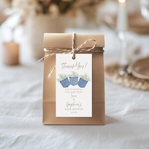 Könnte beinhalten: Eine braune Papiertüte, die mit Bindfaden gebunden ist und ein Etikett mit der Aufschrift "Thank You!" sowie ein Blumendesign mit drei blauen Jeans-Taschen, die mit weißen Blumen gefüllt sind, aufweist. Das Etikett enthält auch den Text "FOR SHOWERING OUR BABY WITH LOVE" und einen Babynamen.