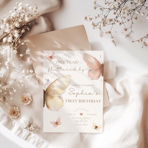 May include: A light beige invitation card with butterfly illustrations and the text "ONE YEAR fluttered by" and "Sophia's FIRST BIRTHDAY". The card includes details for a birthday celebration, with a soft, elegant design.