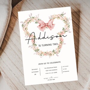 Op de afbeelding: Een roze bloemenkrans met een roze strik in het midden. De tekst "Addison wordt twee" is geschreven in zwarte cursieve letters. De uitnodiging bevat details voor een verjaardagsfeest, inclusief datum, tijd en locatie.
