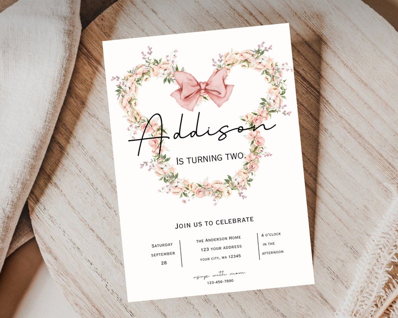 Op de afbeelding: Een roze bloemenkrans met een roze strik in het midden. De tekst "Addison wordt twee" is geschreven in zwarte cursieve letters. De uitnodiging bevat details voor een verjaardagsfeest, inclusief datum, tijd en locatie.
