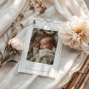 Puede incluir: Una tarjeta blanca con una foto de un bebé durmiendo, enmarcada con un lazo y un borde decorativo. El texto dice "Auntie Amy & Uncle Job" y "Will you be my Godparents?" sobre un fondo neutro con flores.