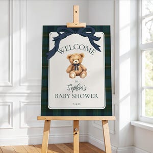 May include: A baby shower welcome sign on an easel. The sign features a teddy bear illustration with a navy bow, the words "WELCOME TO Sophia's BABY SHOWER" and the date 7.14.XX. The background has a navy and green plaid pattern.