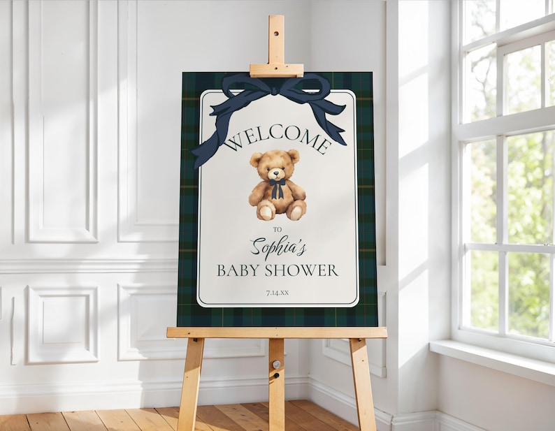 May include: A baby shower welcome sign on an easel. The sign features a teddy bear illustration with a navy bow, the words "WELCOME TO Sophia's BABY SHOWER" and the date 7.14.XX. The background has a navy and green plaid pattern.