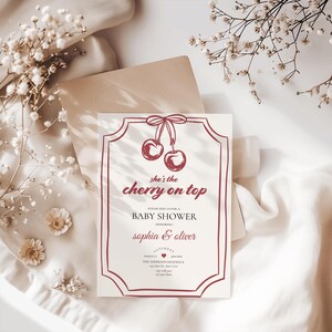 Op de afbeelding: Een rode en witte babyborrel uitnodiging met een kerstillustratie en de tekst "she's the cherry on top". De uitnodiging is voor een babyborrel voor Sophia en Oliver. De datum en locatie van de babyborrel zijn ook inbegrepen.
