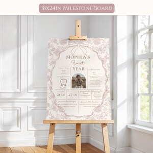 May include: A pink and white milestone board with a floral design and a photo of a baby girl. The board includes information about the baby's first year, including their weight, height, and first tooth. The text on the board reads "Sophia's First Year".