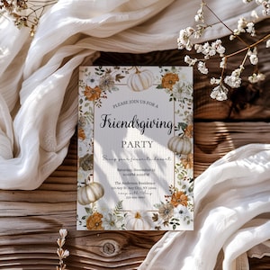 May include: A Friendsgiving party invitation with a floral and pumpkin design. The invitation includes the text "Friendsgiving PARTY" and event details. The invitation is surrounded by white fabric and sits on a wooden surface.