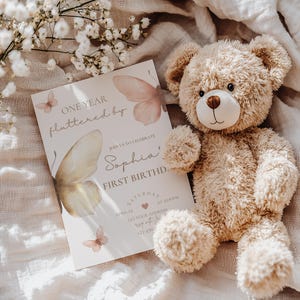 May include: A beige teddy bear next to a first birthday invitation. The invitation features butterfly illustrations and the text "ONE YEAR fluttered by" and "Sophia's FIRST BIRTHD." The invitation also includes the date, time, and address.
