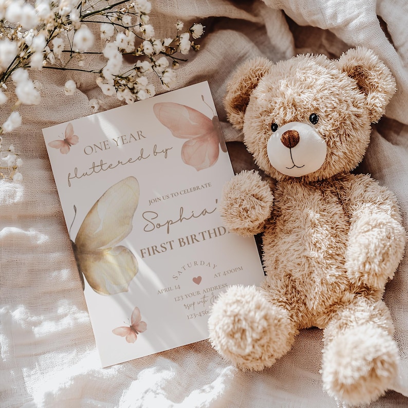May include: A beige teddy bear next to a first birthday invitation. The invitation features butterfly illustrations and the text "ONE YEAR fluttered by" and "Sophia's FIRST BIRTHD." The invitation also includes the date, time, and address.
