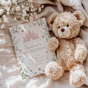 Op de afbeelding: Een roze en witte uitnodigingskaart met een kasteel-illustratie en de tekst "Sophia's First Birthday" op een witte achtergrond met bloemmotieven. De uitnodigingskaart ligt op een wit stoffen oppervlak met een bruine teddybeer.