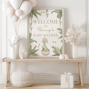 Peut inclure: Une pancarte de bienvenue pour une f&ecirc;te pr&eacute;natale avec un dinosaure vert dans un &oelig;uf, entour&eacute; de feuilles vertes. La pancarte indique "Bienvenue &agrave; la f&ecirc;te pr&eacute;natale de Penny".