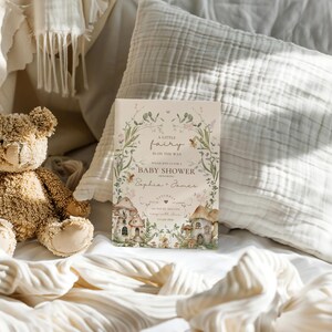 Pu&ograve; includere: Un invito per un baby shower con un tema fatato incantevole. L'invito presenta un bordo floreale con una casa delle fate e il testo "A Little Fairy Is On The Way" e "Baby Shower".