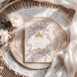 K&ouml;nnte beinhalten: Eine Geburtstagseinladung mit M&auml;rchenmotiv. Sie zeigt ein goldenes Schloss, rosa B&auml;nder und florale Akzente. Der Text lautet "One Upon a Time... Sophia's Third Birthday". Die Einladung liegt auf einem geflochtenen Platzset mit Blumen.