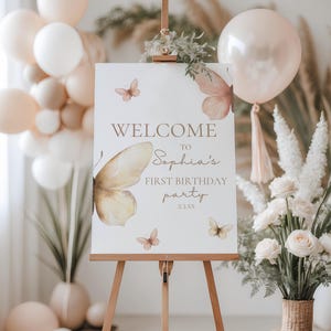 Könnte beinhalten: Eine Holzstaffelei präsentiert ein Willkommensschild für Sophias ersten Geburtstag. Das Schild zeigt Schmetterlingsillustrationen und den Text "WELCOME to Sophia's FIRST BIRTHAY PARTY". Ballons und Blumengestecke in neutralen Tönen umgeben das Schild.