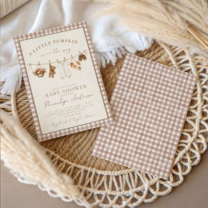 May include: A rectangular baby shower invitation card with a brown gingham border, featuring the text A LITTLE PUMPKIN is on the way, a clothesline illustration with a teddy bear, baby clothes, and autumn leaves, and a matching brown gingham card.
