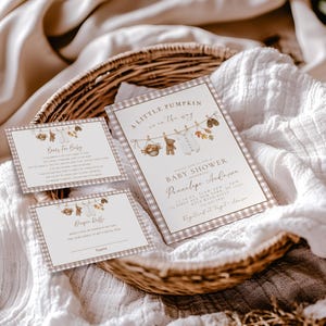 May include: A set of three rectangular baby shower invitation cards with a brown and white gingham border. The cards feature illustrations of a clothesline with a teddy bear, baby clothes, and autumn leaves, with text including A Little Pumpkin.