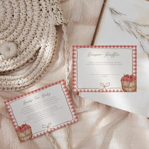 May include: Two rectangular paper cards with a red and white gingham border. One card reads Diaper Raffle with a basket of red apples, and the other reads Books For Baby with a basket of red apples. Both cards feature a small brown line drawing of a bow.