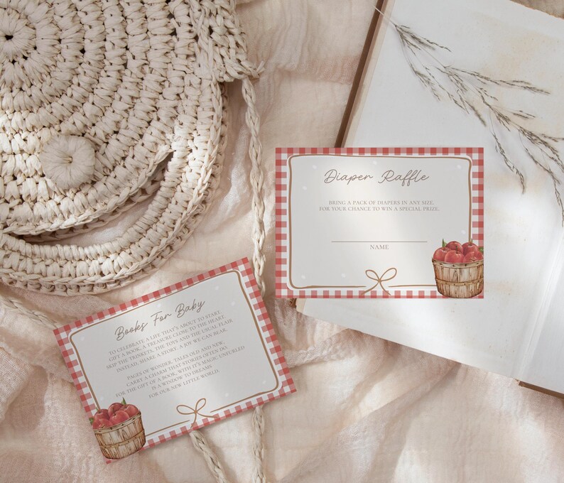 May include: Two rectangular paper cards with a red and white gingham border. One card reads Diaper Raffle with a basket of red apples, and the other reads Books For Baby with a basket of red apples. Both cards feature a small brown line drawing of a bow.