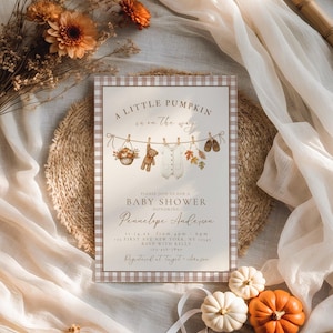 May include: A rectangular baby shower invitation card with a brown and white gingham border. The design features a clothesline with a basket of pumpkins, a teddy bear, a white onesie, autumn leaves, and baby shoes. Text reads A LITTLE PUMPKIN is on the way.
