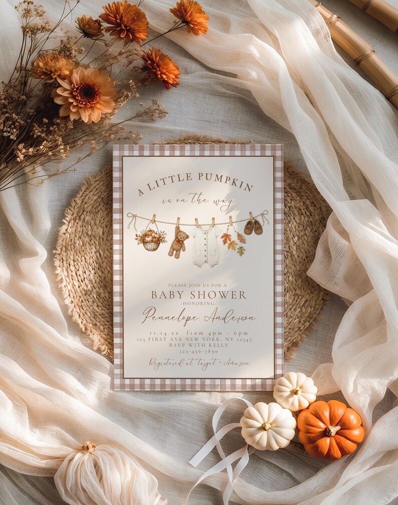 May include: A rectangular baby shower invitation card with a brown and white gingham border. The design features a clothesline with a basket of pumpkins, a teddy bear, a white onesie, autumn leaves, and baby shoes. Text reads A LITTLE PUMPKIN is on the way.
