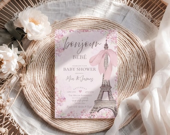 May include: A baby shower invitation with the words "Bonjour Bébé" in script. The invitation features a pink Eiffel Tower design with floral accents. The invitation is on a woven placemat with white flowers and fabric.