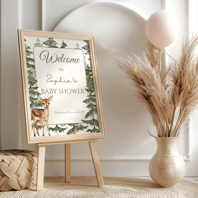 May include: A rectangular sign in a light wood frame on a wooden easel. The sign features a watercolor forest border, a fawn illustration, and text reading Welcome to Sophia's Baby Shower, Saturday December 11th, in dark brown script and print.
