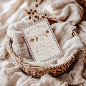 May include: A rectangular baby shower invitation card with a brown and white gingham border. The text reads A LITTLE PUMPKIN is on the way, PLEASE JOIN US FOR A BABY SHOWER HONORING Pennelope Anderson, 12/14 xx from 4pm - 6pm, 123 FIRST AVE NEW YORK, NY 12345, RSVP WITH KELLY 555-555-5555, Registered at Target + Amazon. The card features illustrations of a basket, a teddy bear, a baby onesie, autumn leaves, and shoes hanging on a clothesline.
