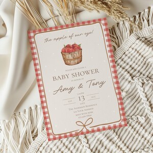 May include: Baby shower invitation with a red gingham border and a watercolor illustration of a basket of apples. The invitation includes the text "The apple of our eye!" and details for Amy & Tony's baby shower.