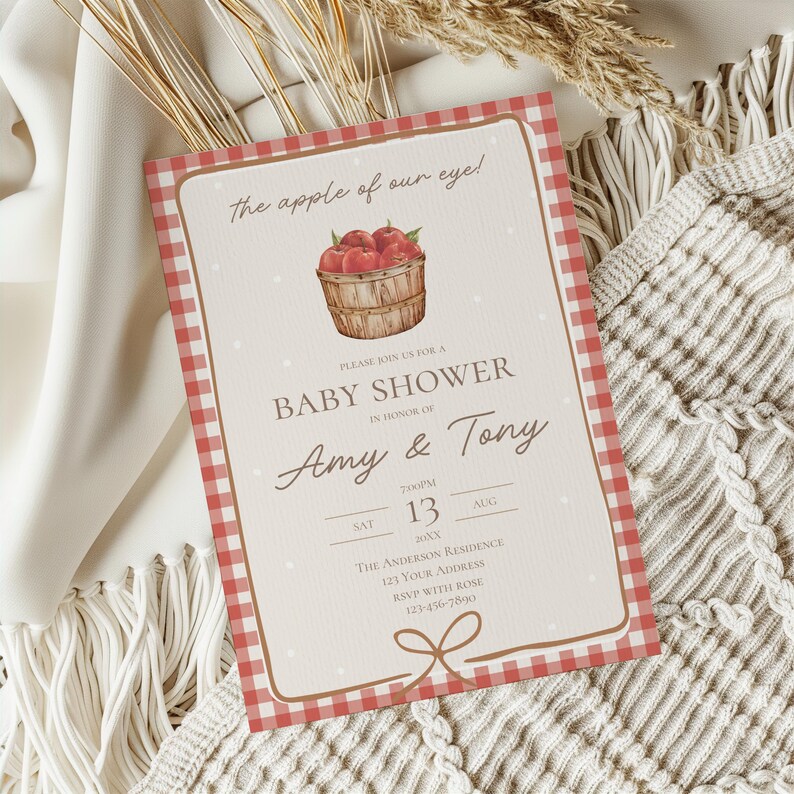 May include: Baby shower invitation with a red gingham border and a watercolor illustration of a basket of apples. The invitation includes the text "The apple of our eye!" and details for Amy & Tony's baby shower.