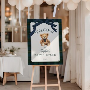 May include: A wooden easel displays a baby shower welcome sign. The sign features a teddy bear illustration, navy blue bow, and text that reads "Welcome to Sophia's Baby Shower". The sign has a plaid border.