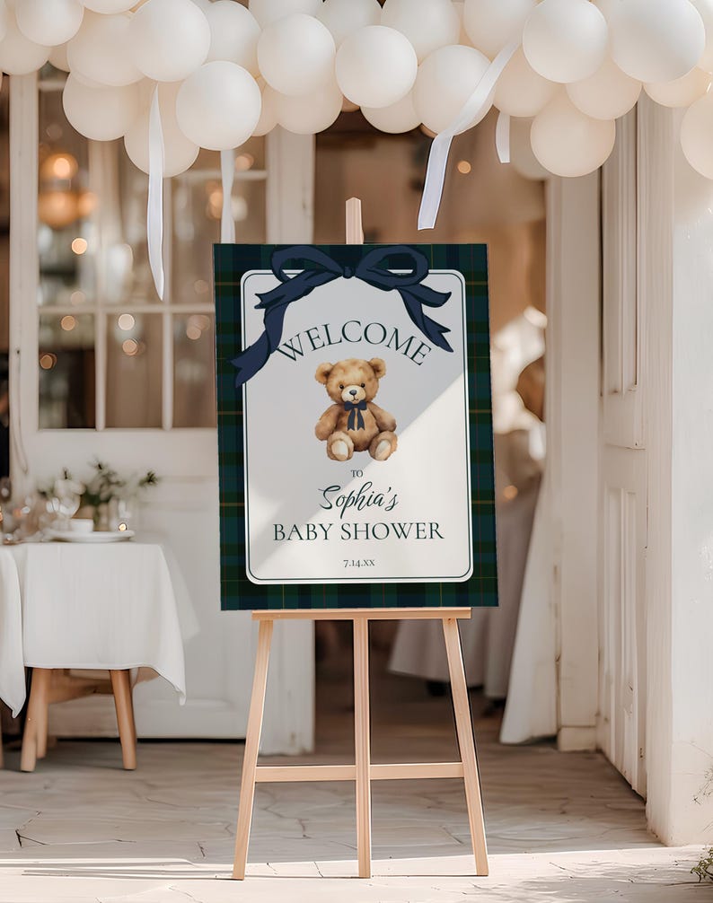 May include: A wooden easel displays a baby shower welcome sign. The sign features a teddy bear illustration, navy blue bow, and text that reads "Welcome to Sophia's Baby Shower". The sign has a plaid border.