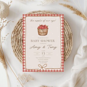 May include: Baby shower invitation with a red and white gingham border. The invitation features the text "The apple of our eye!" and "Baby Shower" with the names "Amy & Tony". A watercolor illustration of a basket of apples is also included.