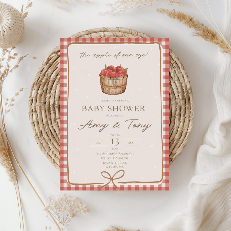 May include: Baby shower invitation with a red and white gingham border. The invitation features the text "The apple of our eye!" and "Baby Shower" with the names "Amy & Tony". A watercolor illustration of a basket of apples is also included.