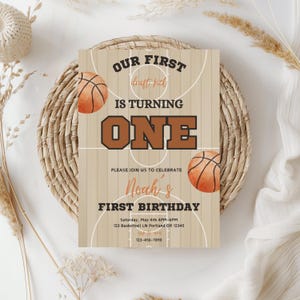 Puede incluir: Una invitación a una fiesta de primer cumpleaños con temática de baloncesto con un fondo de madera marrón. La invitación presenta una cancha de baloncesto con dos balones de baloncesto y el texto "Nuestro primer draft pick cumple un año." La invitación también incluye el nombre "Noah's" y el texto "Primer cumpleaños." La invitación incluye la fecha, la hora y la ubicación de la fiesta.