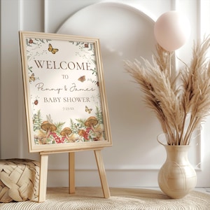 May include: A welcome sign for a baby shower with a woodland theme. The sign is decorated with mushrooms, butterflies, and greenery. The text on the sign reads "Welcome to Penny & James Baby Shower 7-23-XX".