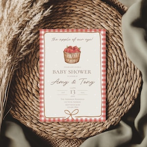 May include: Baby shower invitation with a red and white gingham border. The invitation features the text "The apple of our eye!" and includes a basket of red apples. The invitation is for Amy & Tony.