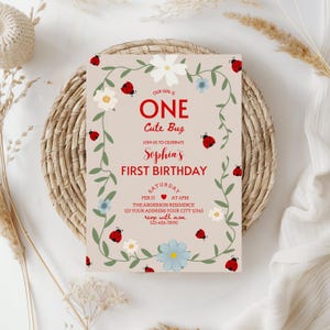 May include: A beige birthday invitation with red text that reads "ONE Cute Bug" and "Sophia's FIRST BIRTHDAY." The invitation is decorated with ladybugs, white and blue flowers, and green vines. The date and time are also included.