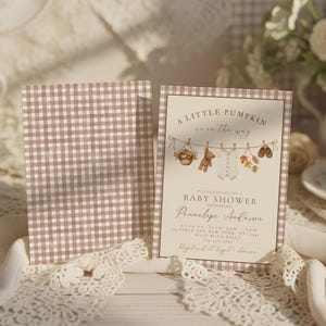 May include: A baby shower invitation card featuring a brown and white gingham border and a central illustration of a clothesline with a teddy bear, baby onesie, and autumn leaves. The text reads A LITTLE PUMPKIN is on the way BABY SHOWER.