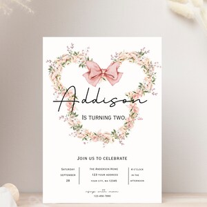 Op de afbeelding: Een roze en witte bloemenkrans met een roze strik in het midden. De tekst "Addison wordt twee" staat onder de krans. De uitnodiging bevat details voor een verjaardagsfeest, inclusief datum, tijd en locatie.