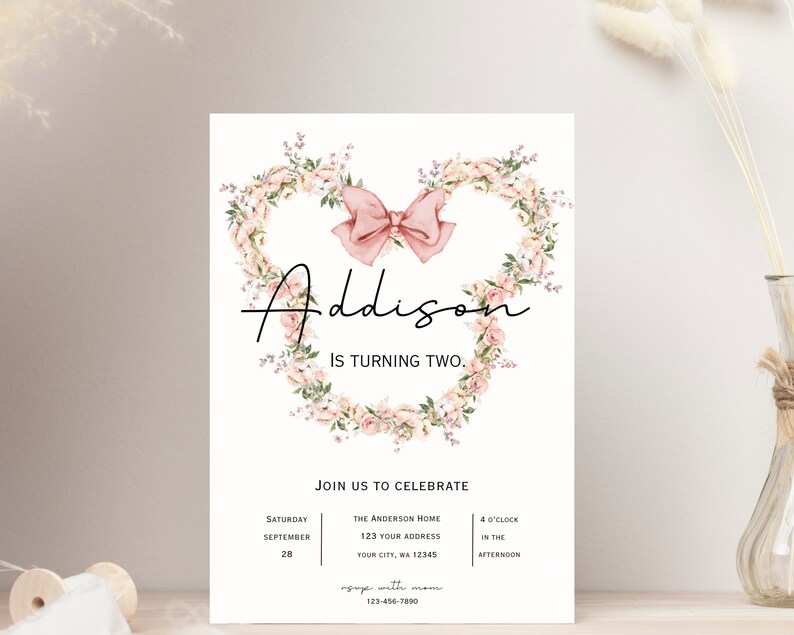 Op de afbeelding: Een roze en witte bloemenkrans met een roze strik in het midden. De tekst "Addison wordt twee" staat onder de krans. De uitnodiging bevat details voor een verjaardagsfeest, inclusief datum, tijd en locatie.