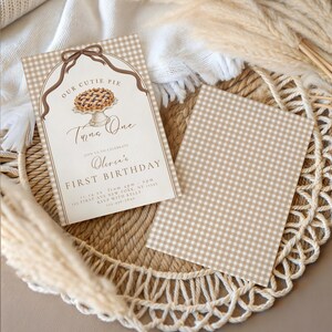 Op de afbeelding: Een rechthoekige uitnodigingskaart met een beige en witte gingham-rand, voorzien van een bruine strik, een taartillustratie en de tekst Our Cutie Pie Turns One Olivia's First Birthday, samen met een bijpassende blanco gingham-kaart.