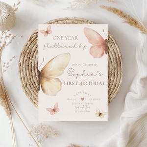 May include: A birthday invitation card with a butterfly theme. The card features the text "ONE YEAR fluttered by" and "Sophia's FIRST BIRTHDAY". The design includes watercolor butterflies in shades of peach and cream, with additional details.
