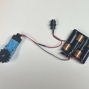 May include: A blue and black electric motor with a gear attached to it. The motor is connected to a battery pack with three AA batteries labeled "DURACELL" and a black wire.