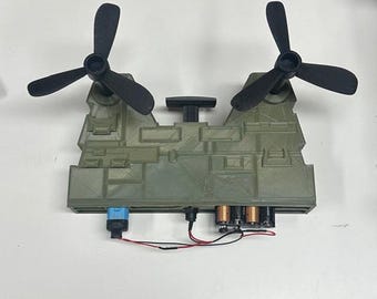 May include: A small, olive green model with two black propellers. The model has a rectangular base with various components, including a blue connector, wires, and cylindrical objects. The propellers are positioned on top of the model.
