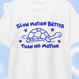 Peut inclure: Un t-shirt blanc à manches courtes avec un motif bleu représentant une tortue et le texte "SLOW MOTION BETTER THAN NO MOTION" accompagné de petites étoiles. Le vêtement est présenté sur un cintre en bois sur fond bleu clair.