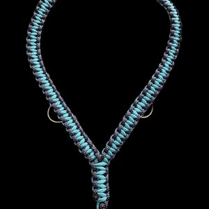 May include: A Y-shaped harness constructed from braided paracord in alternating light blue and dark blue with speckled patterns. The harness features three circular metal rings positioned at the sides and bottom, secured with a black plastic buckle.
