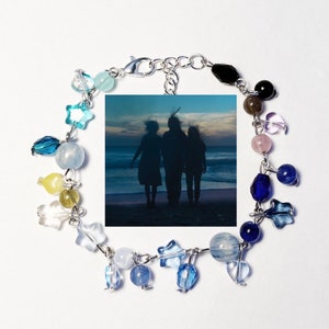 Pode incluir: Um bracelete de prata com uma variedade de contas de vidro azul, amarelo e transparente, pingentes em forma de estrela e um pingente retangular com foto que mostra a silhueta de três pessoas numa praia.