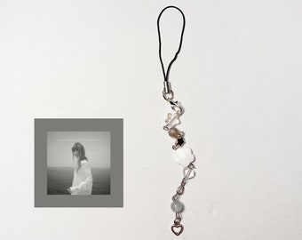 May include: A white and black charm phone strap with a heart, beads, and flowers. The strap is attached to a black cord.