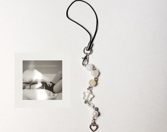May include: A white and yellow beaded phone charm with a silver heart, star, and beads. The charm is attached to a black string.