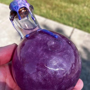 May include: A round glass bottle filled with a shimmering purple liquid. The bottle has a cork stopper with blue and purple dripped accents. The bottle is held in a hand, with a blurred background of green grass and a sidewalk.